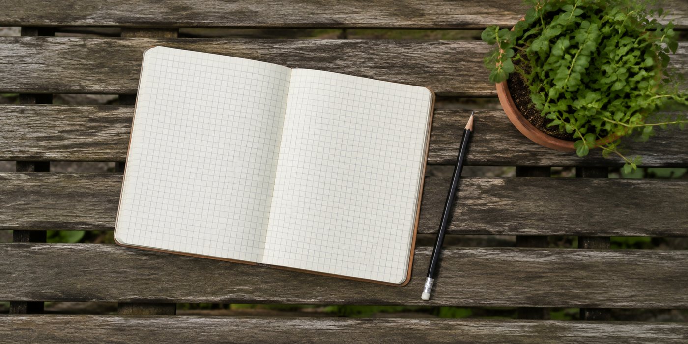 Caderno de folha quadriculada aberto e um l&aacute;pis pousados num banco de jardim de madeira, ao lado de um vaso de ervas